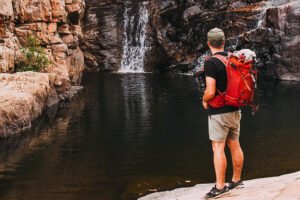 There is a man standing near the waterfall with a red rucksack.