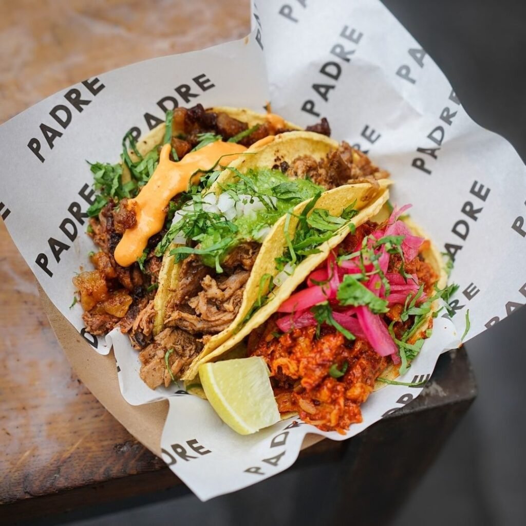 Three soft tacos filled with slow-cooked meats, fresh cilantro, pickled onions, and a drizzle of orange sauce, served on branded Padre paper with a lime wedge at Tacos Padre, London