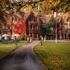 A beautiful autumn view of a grand building in the Cotswolds, covered in red ivy. The scenic pathway leads up to the entrance, surrounded by lush greenery and trees with golden foliage.