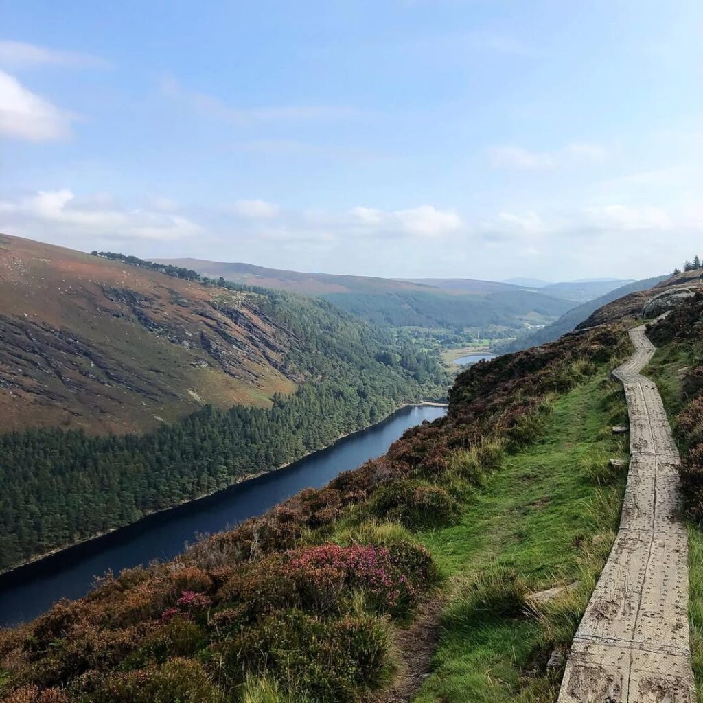 The Spinc trail in Glendalough, County Wicklow, Ireland, featuring a scenic boardwalk path along rugged hillsides, overlooking lush valleys and tranquil lakes under a clear blue sky