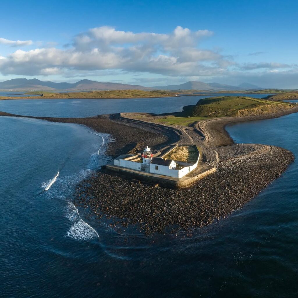 Inishgort Lighthouse in Clew Bay, County Mayo, surrounded by calm waters and dramatic drumlin islands, with a backdrop of rolling hills and a bright blue sk