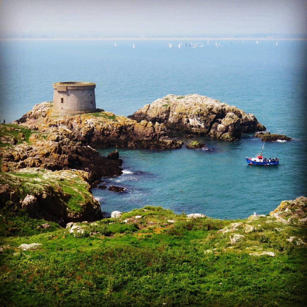 Ireland's Eye, a picturesque island off the coast of Howth, featuring a historic Martello Tower, rugged cliffs, and turquoise waters, with a vibrant fishing boat adding charm to the serene scene