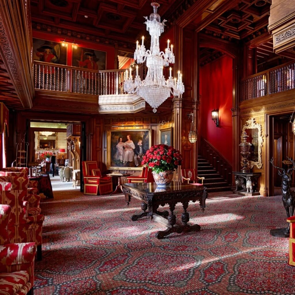 Majestic interior of Ashford Castle, featuring rich wooden paneling, a sparkling crystal chandelier, intricate artwork, and lavish red furnishings exuding timeless elegance