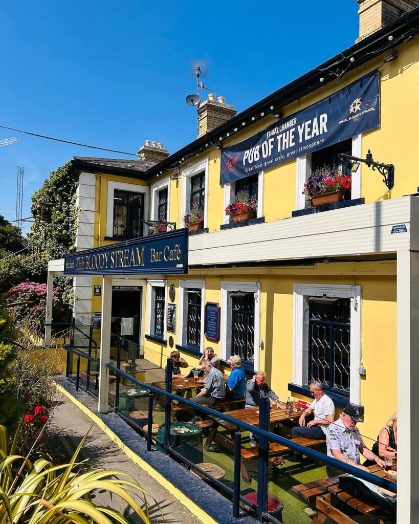 The Bloody Stream Bar & Café in Howth, a charming yellow pub with outdoor seating, vibrant flower displays, and a relaxed atmosphere, perfect for enjoying sunny days and great food