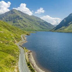 Scenic winding road along a tranquil lake in County Mayo, surrounded by lush green mountains under a clear blue sky.