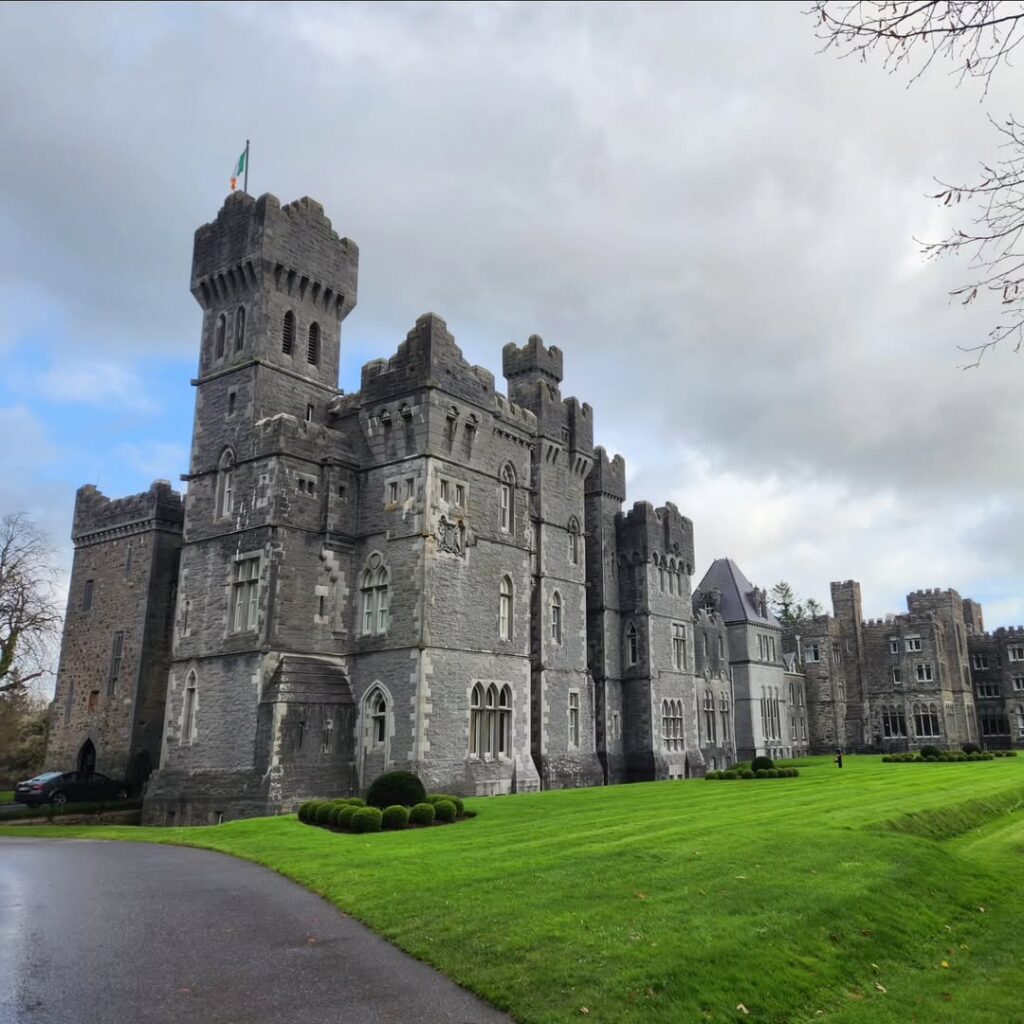Ashford Castle, an 800-year-old medieval castle in County Mayo, Ireland, featuring stunning stone architecture, lush green lawns, and a majestic presence under a cloudy sky