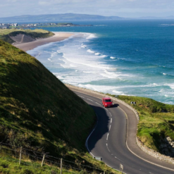 Scenic coastal road in Ireland with a red car driving along the shoreline, overlooking rolling waves and sandy beaches under a clear blue sky.