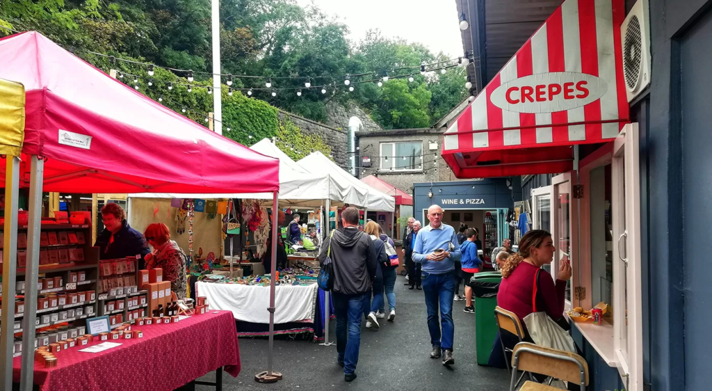 A vibrant scene at Howth Market, featuring colorful stalls, local crafts, food vendors, and visitors enjoying the lively atmosphere under a cozy, charming setting