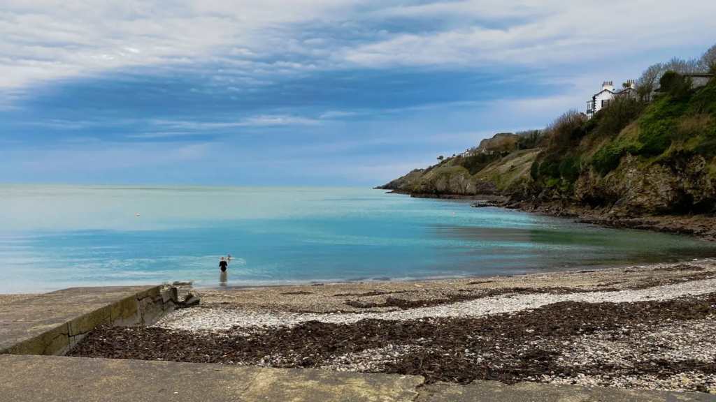 Sutton Strand beach is one of the best things to do in howth with its turquoise waters, rocky cliffs, and a lone swimmer under a cloudy sky.
