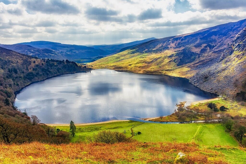 Stunning view of Lough Tay, also known as the Guinness Lake, in County Wicklow, Ireland, surrounded by vibrant green hills, rugged cliffs, and illuminated by soft sunlight under a partly cloudy sky.