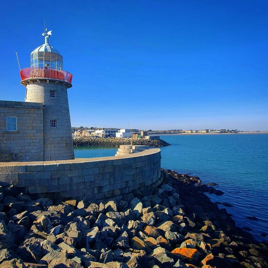The Howth Lighthouse standing gracefully at the edge of the pier, surrounded by calm blue waters and a clear sky, offering stunning views of Dublin Bay