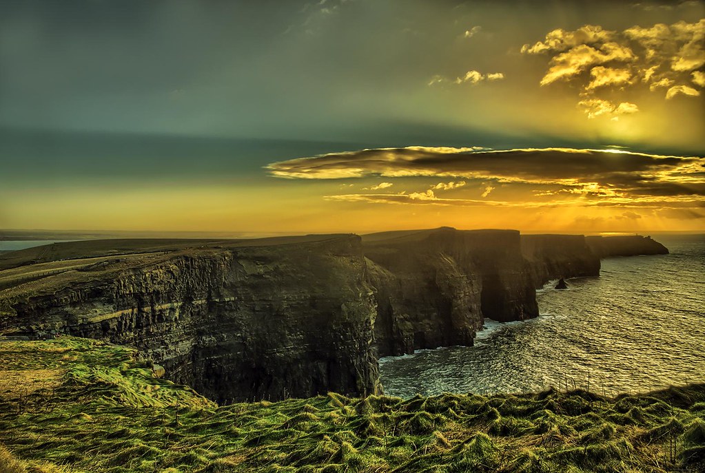 A stunning sunset view over the Cliffs of Moher, with the cliffs towering above the Atlantic Ocean and the sky painted with golden and orange hues.