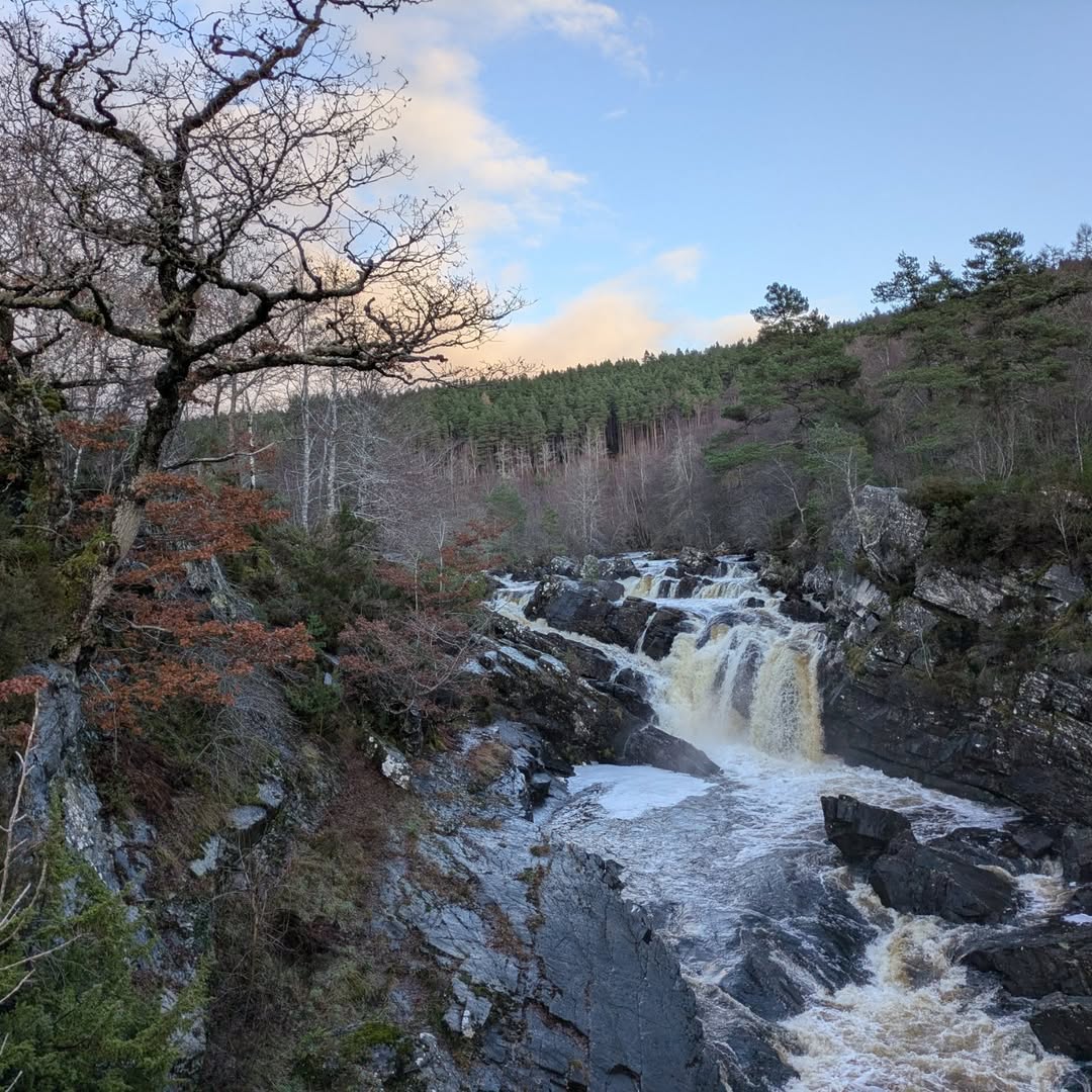 Cascading waterfall surrounded by lush trees and rocky terrain along the North Coast 500 Scotland under a pastel evening sky.