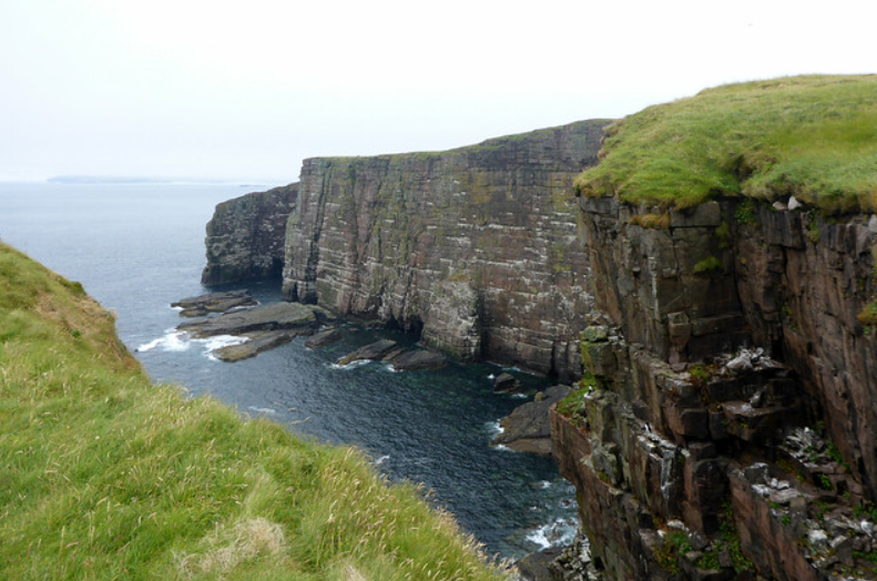 Dramatic coastal cliffs with sheer drops into the sea, covered in grassy edges under a cloudy sky.
