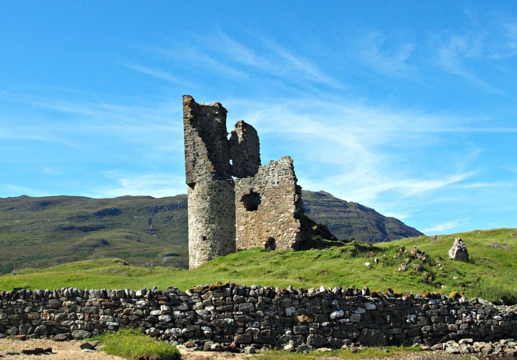 Ruins of Ardvreck Castle on a grassy hill with mountains in the background along the North Coast 500 Scotland under a bright blue sky.