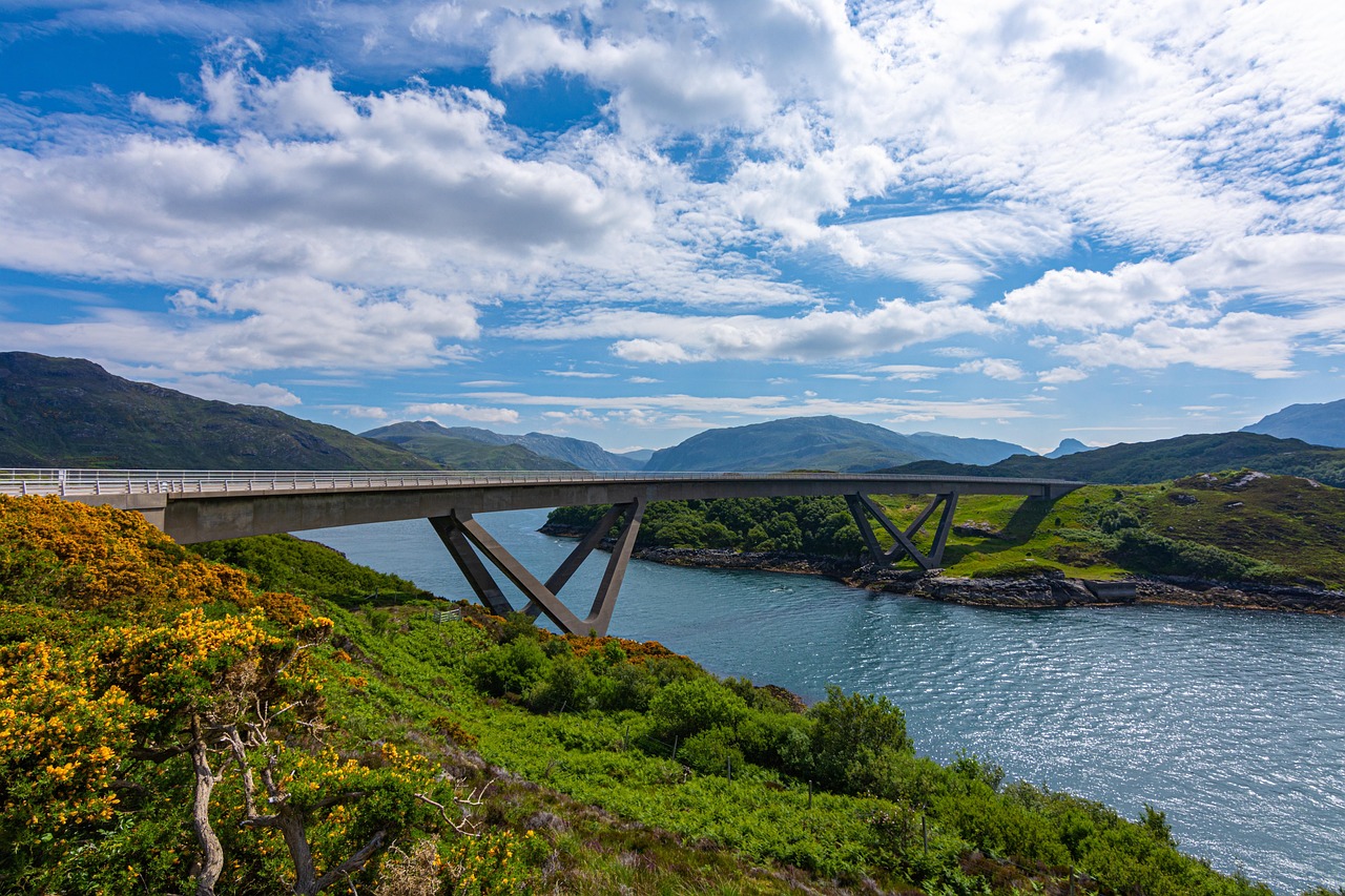 Scenic view of Kylesku Bridge along the North Coast 500 Scotland, surrounded by water, green hills, and mountains under a blue sky.