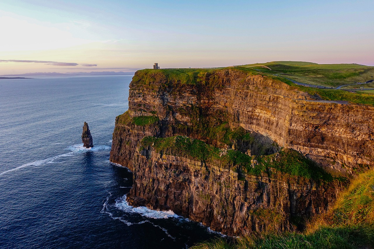 A scenic view of the Cliffs of Moher in Ireland at sunset, featuring rugged cliffs with patches of green grass, a lone sea stack in the ocean, and O'Brien's Tower perched on the cliff edge under a pastel sky.