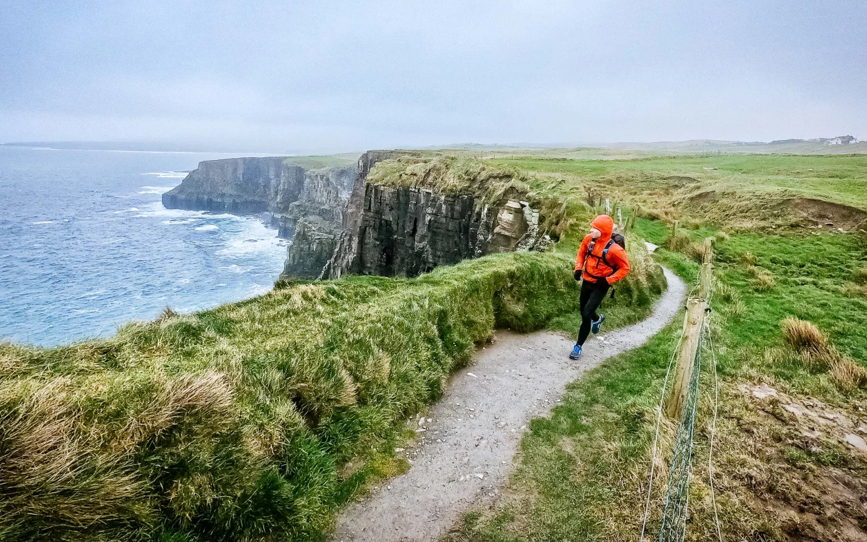 A person jogging along the scenic trail at the Cliffs of Moher, with the dramatic cliffs and the Atlantic Ocean visible in the background