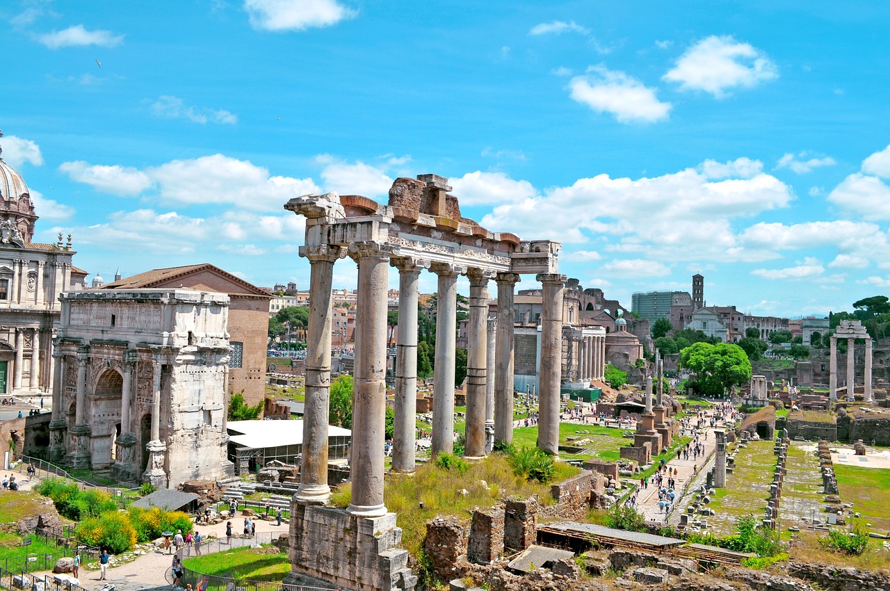 Ruins of the Roman Forum in Rome, Italy, featuring ancient columns, arches, and historic structures under a bright blue sky with scattered clouds.