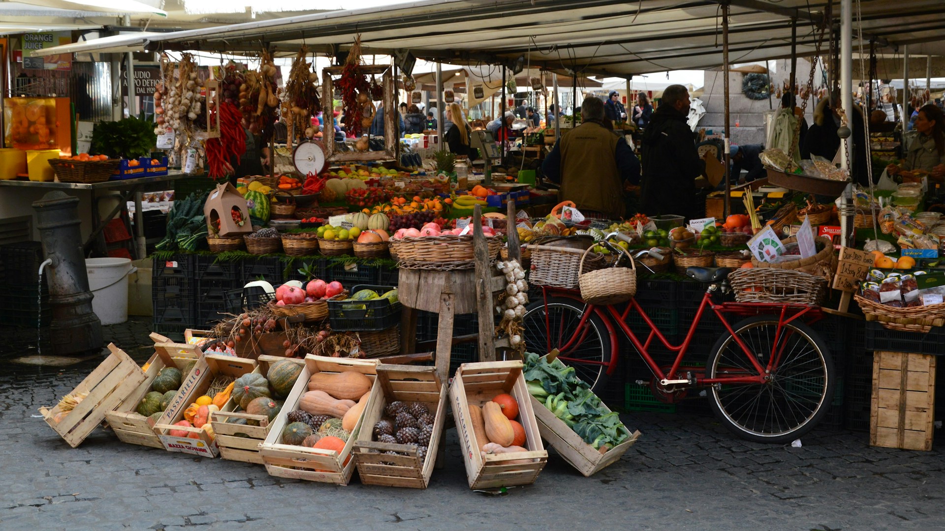 A vibrant outdoor market in Rome with fresh fruits, vegetables, and spices displayed in wooden crates, alongside a red bicycle.