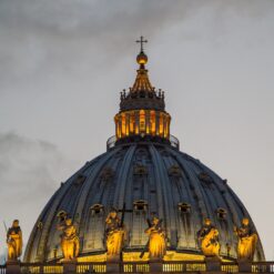Illuminated dome of St. Peter's Basilica in Vatican City at dusk, showcasing statues and intricate architecture.